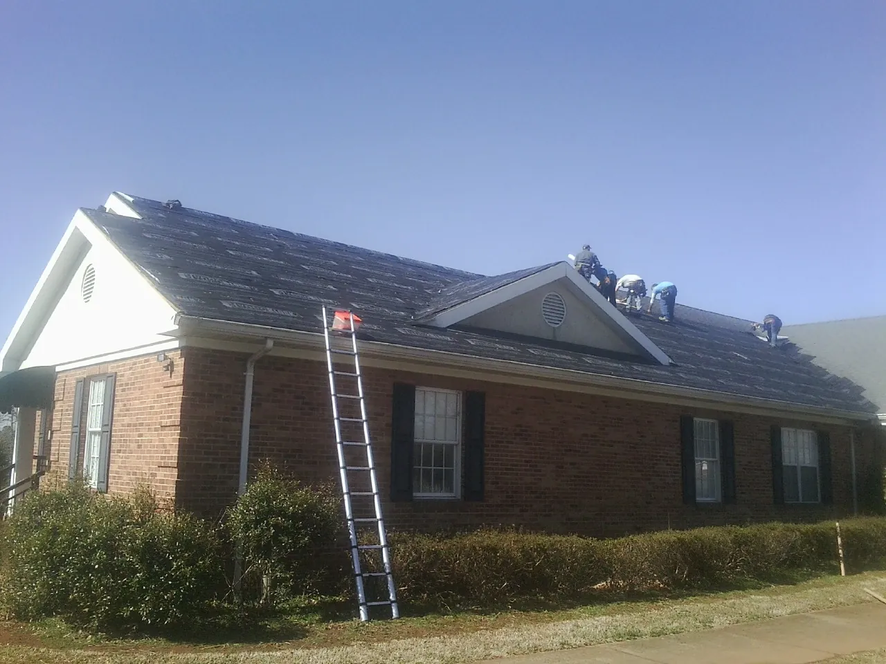 Crew tearing shingles off a brick ranch roof with a ladder set — Emergency Roof Repair work by our crew in Roslyn Heights