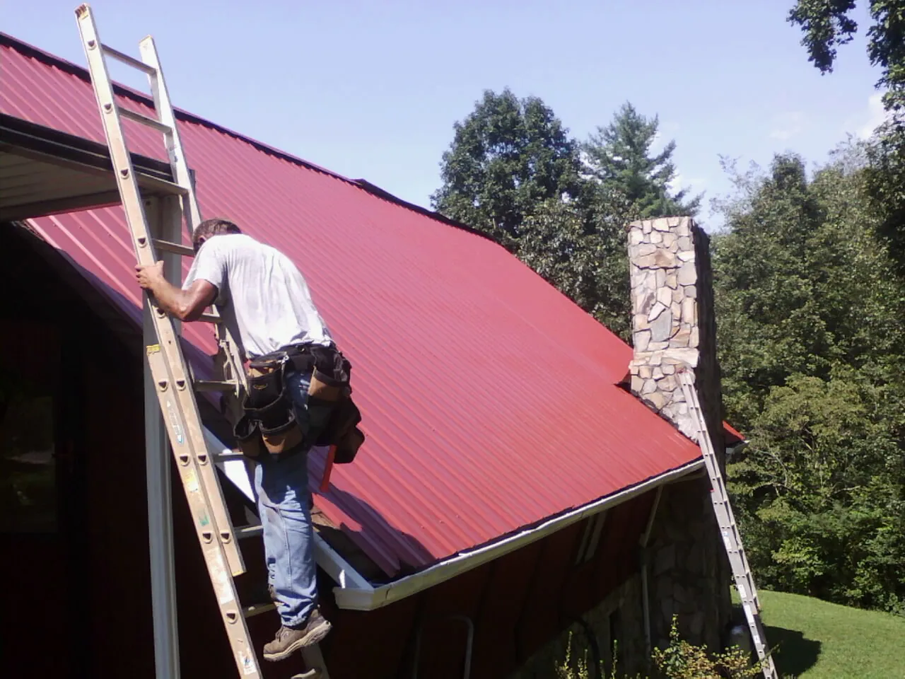 Roofer on a ladder beside a red metal roof and stone chimney — Soffit & Fascia Repair by Roofing Brothers in Roslyn Heights