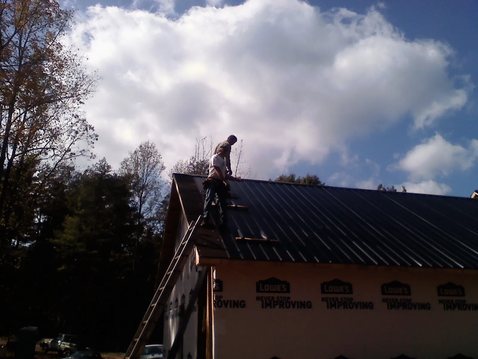 Roofer fastening dark metal panels high against a cloudy sky, from a Roof Inspection job in Roslyn Heights